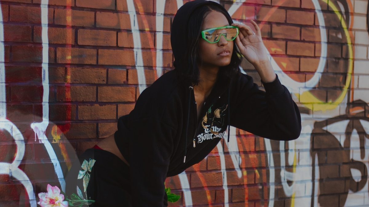 Model in a black cropped hoodie and green sunglasses against a graffiti brick wall